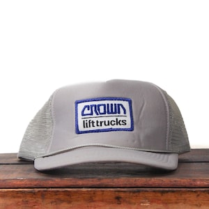 May include: A gray trucker hat with a mesh back and a curved brim. The front features a rectangular patch with the words "CROWN lift trucks" in white and blue lettering.