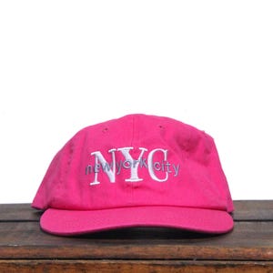 Puede incluir: Gorra de béisbol rosa brillante con el texto bordado "NYC new york city" en blanco. La gorra está hecha de una tela suave y tiene una visera curva. El sombrero está sobre una superficie de madera.