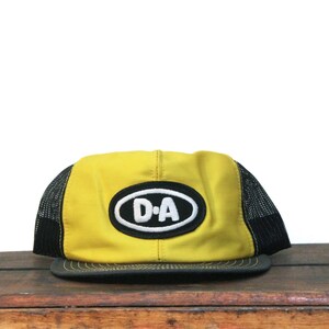 May include: Yellow and black trucker hat with a black brim. The hat features a white oval patch with the letters "D-A" in black. The hat is sitting on a wooden surface.