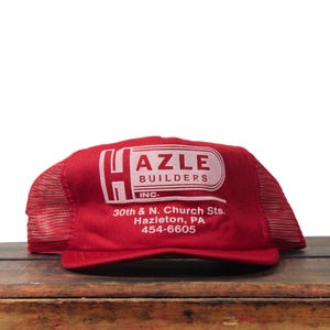 Puede incluir: Gorra de camionero roja con el logotipo blanco "HAZLE BUILDERS INC.". La gorra tiene una parte trasera de malla y una visera curva. El texto "30th & N. Church Sts. Hazleton, PA 454-6605" está impreso debajo.