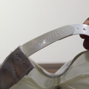 May include: Close-up of a white baseball cap's adjustable strap. The strap is made of plastic and features the embossed text "MADE IN USA". The cap is off-white and has a snap closure.