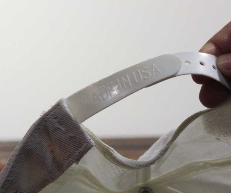 May include: Close-up of a white baseball cap's adjustable strap. The strap is made of plastic and features the embossed text "MADE IN USA". The cap is off-white and has a snap closure.