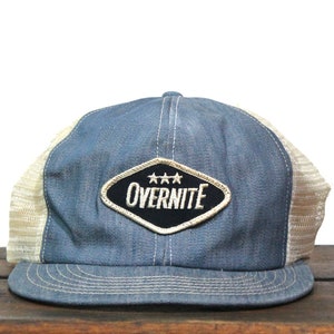 Puede incluir: Una gorra de camionero de mezclilla azul con una malla blanca en la parte posterior. La gorra tiene un parche en forma de diamante con el texto "OVERNITE" en letras blancas y tres estrellas sobre el texto.