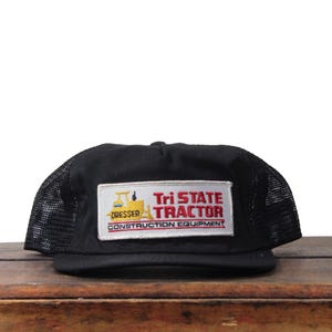 May include: Black trucker hat with a mesh back and a flat brim. The hat features a rectangular patch with the text "Tri State Tractor CONSTRUCTION EQUIPMENT" in red and white, with a yellow tractor graphic.