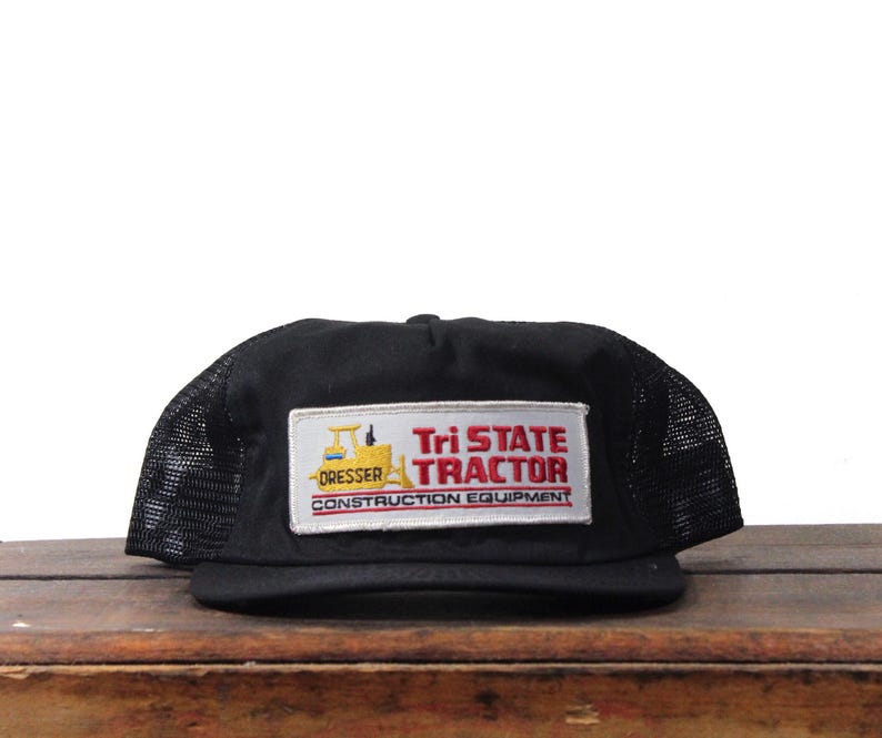 May include: Black trucker hat with a mesh back and a flat brim. The hat features a rectangular patch with the text "Tri State Tractor CONSTRUCTION EQUIPMENT" in red and white, with a yellow tractor graphic.