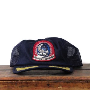 May include: Navy blue trucker hat with a mesh back and a curved brim. The front features a patch with the text "Columbia Spacelab 1" and an illustration of a spacecraft and the moon. The brim has gold embroidery.