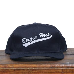 Może przedstawiać: Czarna czapka bejsbolówka z białym zakrzywionym logo z napisem "Berger Bros."