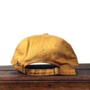 K&ouml;nnte beinhalten: Senffarbene Baseballkappe mit gebogenem Schirm und verstellbarem Riemen. Die Kappe hat ein kleines, schwarzes gesticktes Design auf der Seite. Die R&uuml;ckseite der Kappe hat eine Metallschnalle zur Einstellung.