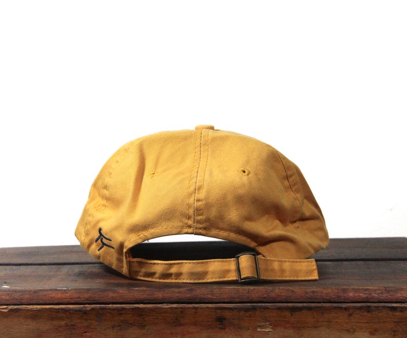 K&ouml;nnte beinhalten: Senffarbene Baseballkappe mit gebogenem Schirm und verstellbarem Riemen. Die Kappe hat ein kleines, schwarzes gesticktes Design auf der Seite. Die R&uuml;ckseite der Kappe hat eine Metallschnalle zur Einstellung.