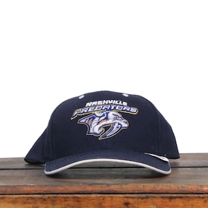 Może przedstawiać: Czapka bejsbolowa w kolorze granatowym z białym daszkiem. Z przodu czapki znajduje się haftowane logo drużyny hokejowej Nashville Predators. Logo jest żółte, niebieskie i białe i przedstawia stylizowanego drapieżnika z nazwą drużyny.