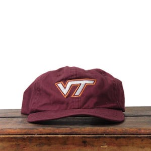Op de afbeelding: Bordeauxrode baseballcap met een wit en oranje "VT" logo. De cap is gemaakt van een zacht materiaal en heeft een gebogen klep. De hoed ligt op een houten oppervlak.