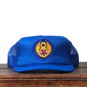 Puede incluir: Gorra de camionero azul con parte trasera de malla y visera curva. La parte delantera de la gorra presenta un parche circular con un emblema dorado y una estrella roja y blanca. La gorra está sobre una superficie de madera.