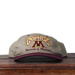 May include: A khaki baseball cap featuring the word "Gophers" in gold and a maroon "M" logo. The brim is maroon, with "University of Minnesota" embroidered below. The cap is displayed on a wooden surface.