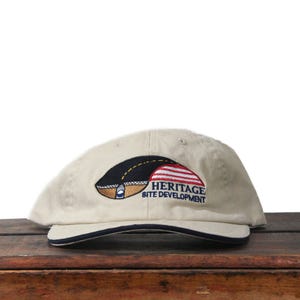 May include: A beige baseball cap with a navy blue trim, displaying the text "HERITAGE SITE DEVELOPMENT." The logo incorporates a black and gold design alongside a red and white striped element.