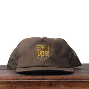 Può includere: Cappellino da camionista marrone con retro in rete e visiera curva. La parte anteriore del cappellino presenta il logo UPS ricamato in oro. Il cappellino è su una superficie di legno.