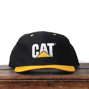 May include: Black baseball cap with a yellow brim. The cap features the white embroidered letters "CAT" above a yellow and white logo.