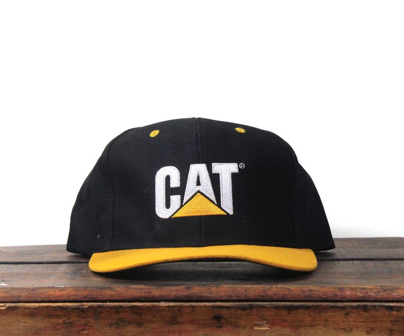 May include: Black baseball cap with a yellow brim. The cap features the white embroidered letters "CAT" above a yellow and white logo.
