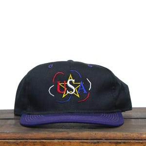 May include: A black baseball cap with a purple peak. The cap has the embroidered word "USA" in red, white, and blue, with a yellow star and a white outline. The cap is on a wooden surface.