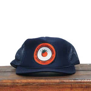 May include: A navy blue trucker hat with a mesh back and a curved brim. The hat features a circular patch with a tomato graphic and the text "BOARD OF COUNTY COMMISSIONERS ORANGE COUNTY FLORIDA".