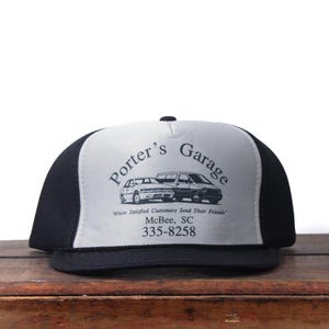 May include: A black and white trucker cap featuring the text "Porter's Garage" and an image of two cars. The cap also includes the text "Where Satisfied Customers Send Their Friends" and a phone number.
