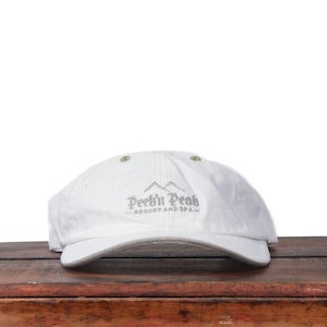 May include: A white baseball cap with the text "Peek'n Peak Resort and Spa" in gray script. The cap has a curved brim and a button on top. The hat is sitting on a wooden surface.