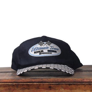 May include: A black baseball cap with a checkered brim. The cap features an embroidered oval logo with the words "Ultimate Team," "Kohler," and "Ferguson." The logo includes two checkered flags and a light blue accent.