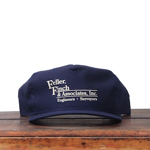 Könnte beinhalten: Marineblaue Baseballkappe mit flachem Schirm. Auf der Kappe steht in weißer Schrift "Feller, Finch & Associates, Inc. Engineers • Surveyors."