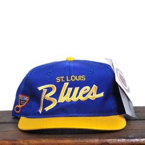 Op de afbeelding: Een blauwe en gele baseballpet met de tekst "St. Louis Blues" geborduurd op de voorkant. De pet heeft een gele klep en een blauwe kroon.