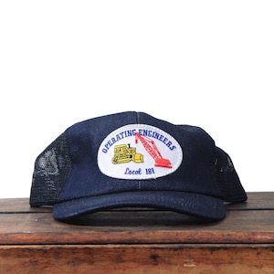 Può includere: Un cappellino da camionista blu navy con retro in rete e visiera curva. Il cappello presenta una toppa con le parole "Operating Engineers" e un logo di un bulldozer e una gru. Il testo "Local 181" è anche sulla toppa.
