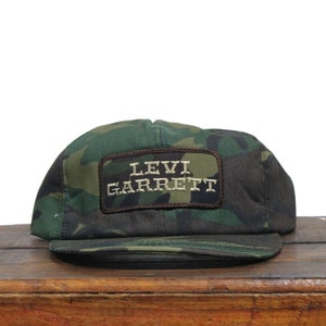 May include: A camouflage green and brown baseball cap with a brown patch that reads "Levi Garrett" in white embroidered letters.