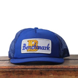 Puede incluir: Gorra de camionero azul con parte trasera de malla y visera curva. La parte delantera presenta un parche rectangular con la palabra "Benchmark" en escritura azul y una "B" dorada.