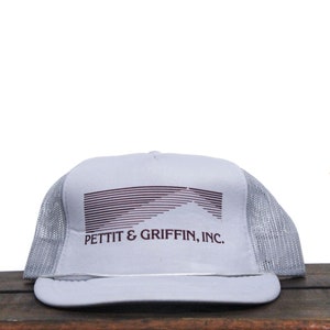 Puede incluir: Una gorra de camionero gris y blanca con un logotipo burdeos que dice "Pettit & Griffin, Inc.". El logotipo presenta un diseño de onda estilizado.
