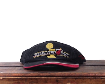 May include: Black visor with the text "2006 NBA CHAMPIONS" embroidered in gold and red. The visor features a gold trophy and the Miami Heat logo. The brim has a red and white stripe.