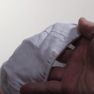 Pu&ograve; includere: Un cappellino da baseball bianco con visiera curva. Il cappellino &egrave; tenuto in mano, mostrando il lato inferiore della visiera e le cuciture interne. Ci sono alcune piccole macchie sul cappellino.