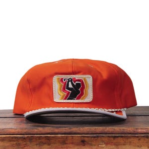 Può includere: Cappellino da baseball arancione con visiera bianca e finiture bianche decorative. Il cappellino presenta una toppa con una silhouette di golfista in nero, con una mazza da golf, su uno sfondo giallo e rosso. Il cappellino è appoggiato su una superficie di legno.