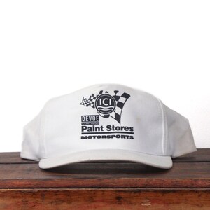 May include: White baseball cap with a curved brim. The cap features black text and graphics, including the words "DEVOE COATINGS PAINT STORES MOTORSPORTS" and a checkered flag design. The cap is sitting on a wooden surface.