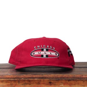 Puede incluir: Gorra de béisbol roja con el texto "CHICAGO BULLS" bordado en la parte delantera. La gorra tiene una visera plana y una corona estructurada. El logotipo es un óvalo con rayas blancas y negras.