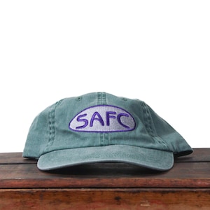 Könnte beinhalten: Eine teal Baseballkappe mit den Buchstaben "SAFC" in Lila in einem Oval gestickt. Die Kappe besteht aus einem weichen Material und liegt auf einer Holzoberfläche. Die Kappe hat einen gebogenen Schirm.