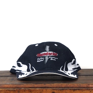 Może przedstawiać: Czarna czapka bejsbolowa z białymi płomieniami po bokach i na rondzie. Czapka ma napis "Thunder Jet" w kolorze czerwonym i białym, z dodatkowym tekstem poniżej. Kapelusz leży na drewnianej powierzchni.