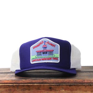 Könnte beinhalten: Blau-weiße Trucker-Mütze mit einem Aufnäher mit einem Gebäude, einer Flagge und dem Text "Springfield Illinois Lincoln Heritage Trail". Die Mütze hat einen Mesh-Rücken und einen gebogenen Schirm.