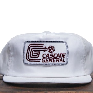 May include: White trucker hat with a burgundy and silver embroidered patch that says "CASCADE GENERAL" with a stylized "G" and a soccer ball graphic.