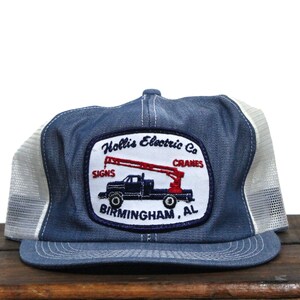 Puede incluir: Gorra de camionero azul y blanca con un parche que muestra un camión con una grúa y el texto "Hollis Electric Co. Signs Cranes Birmingham, AL."