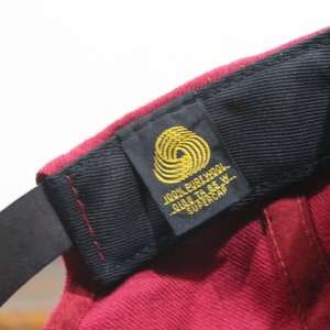 May include: A burgundy wool baseball cap with a black strap and a gold label that reads "100% PURE WOOL SIZE TO 6 5/8  SUPERCAP"