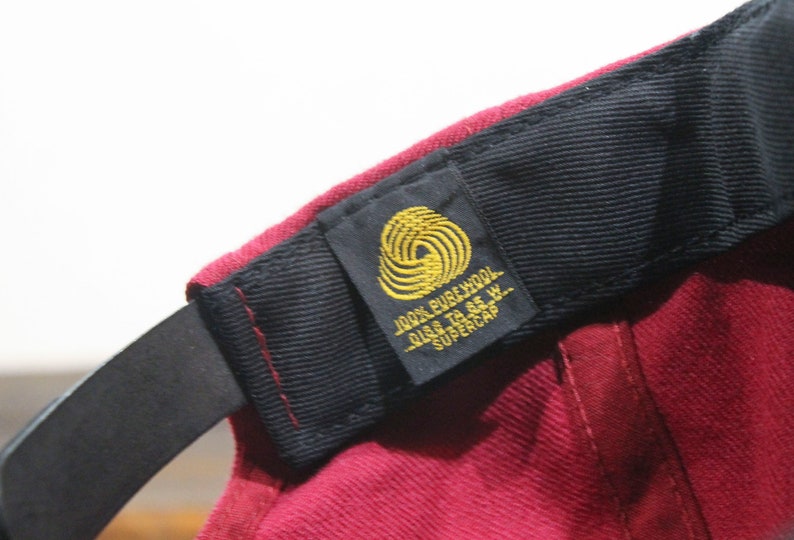 May include: A burgundy wool baseball cap with a black strap and a gold label that reads "100% PURE WOOL SIZE TO 6 5/8  SUPERCAP"
