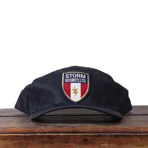 May include: Black baseball cap with a curved peak. The cap has a patch with the text "STORM SECURITY, LTD." above a red, white, and gold shield with a lightning bolt. The cap is on a wooden surface.