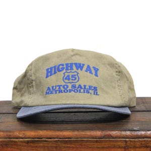 May include: A tan and blue baseball cap with the text "HIGHWAY 45 AUTO SALES METROPOLIS, IL" in blue.