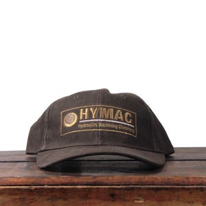 May include: A dark brown baseball cap with the text "HYMAC" in gold lettering. The cap has a curved brim and is sitting on a wooden surface. The text "Hydraulics Machining Chroming" is below the main text.