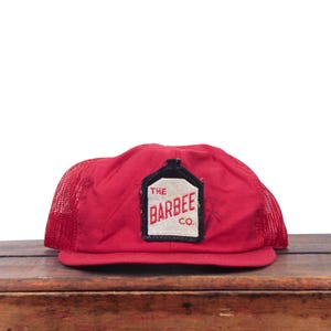 May include: A red trucker cap featuring a mesh back and a solid front panel. A rectangular patch is centred on the front, displaying the text THE BARBEE CO. in red lettering on a white background with a black border shaped like a bottle.