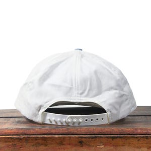 May include: A white baseball cap with a curved brim and adjustable snapback closure. The cap has a light blue top button and a white strap. The hat is made of a soft, textured material.