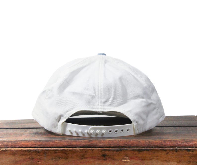 May include: A white baseball cap with a curved brim and adjustable snapback closure. The cap has a light blue top button and a white strap. The hat is made of a soft, textured material.